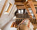 A cozy loft area in a wooden cabin features exposed beams, a staircase, natural wood finishes, and windows letting in sunlight. A patterned blanket is draped over a chair in the foreground.