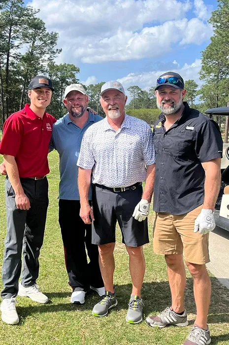 Four men in golf attire stand together outdoors on a sunny day, smiling at the camera. Trees and a golf cart are visible in the background, suggesting they are on a golf course.