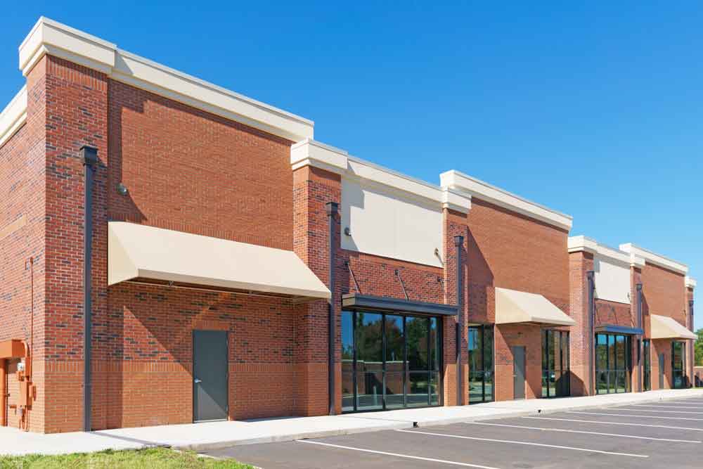 A row of empty brick retail stores with large windows and beige awnings, set in front of an empty parking lot under a clear blue sky.