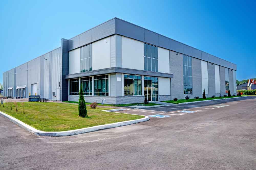 A large, modern industrial warehouse with glass windows, grey walls, and a flat roof, surrounded by a parking lot and landscaped grass on a clear, sunny day.