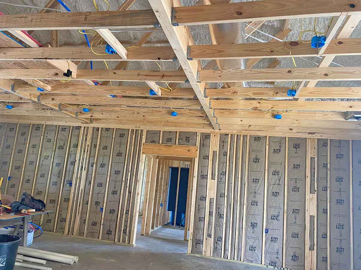 Wood framing and insulation are visible inside a building under construction. Exposed beams and electrical wiring run across the ceiling, while insulated walls line the room. Construction tools and materials are scattered on the floor.