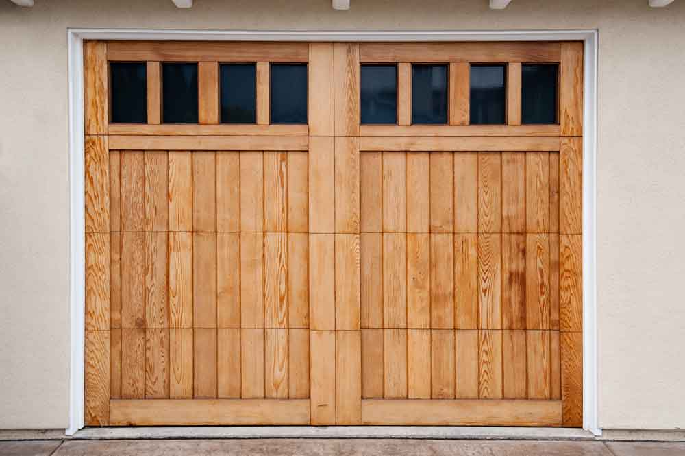 A wooden double garage door with vertical panels and six small, rectangular windows at the top. The door is set in a light-colored wall with a white trim around the edges.