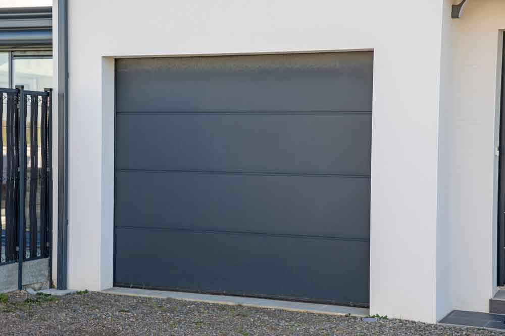 A closed modern gray garage door set in a white exterior wall, with part of a glass structure and metal railing visible on the left and a small section of a doorway on the right. Gravel covers the ground in front.