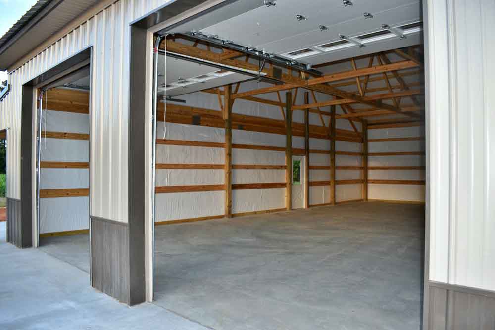 Large empty metal garage with open doors, showing a spacious interior with exposed wooden beams, metal siding, and concrete floor—an ideal space for adding garage insulation. Daylight streams through the openings and a small side window.