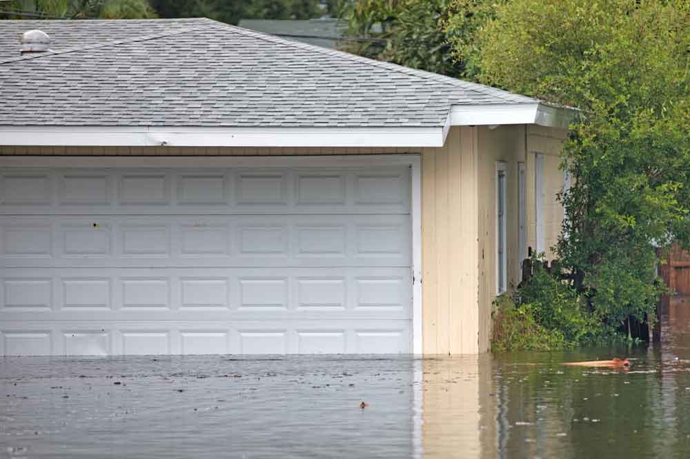 A residential garage with white doors is partially submerged in floodwater, with water reaching halfway up the doors. Green bushes are visible to the right of the garage.