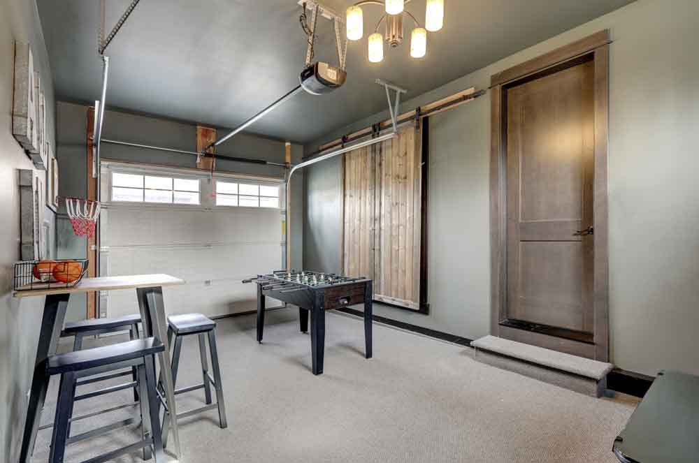 A small converted garage features a foosball table, a tall table with stools, a mini basketball hoop, carpeted floor, garage door, and a wooden door leading into the house. Ceiling lights hang overhead.