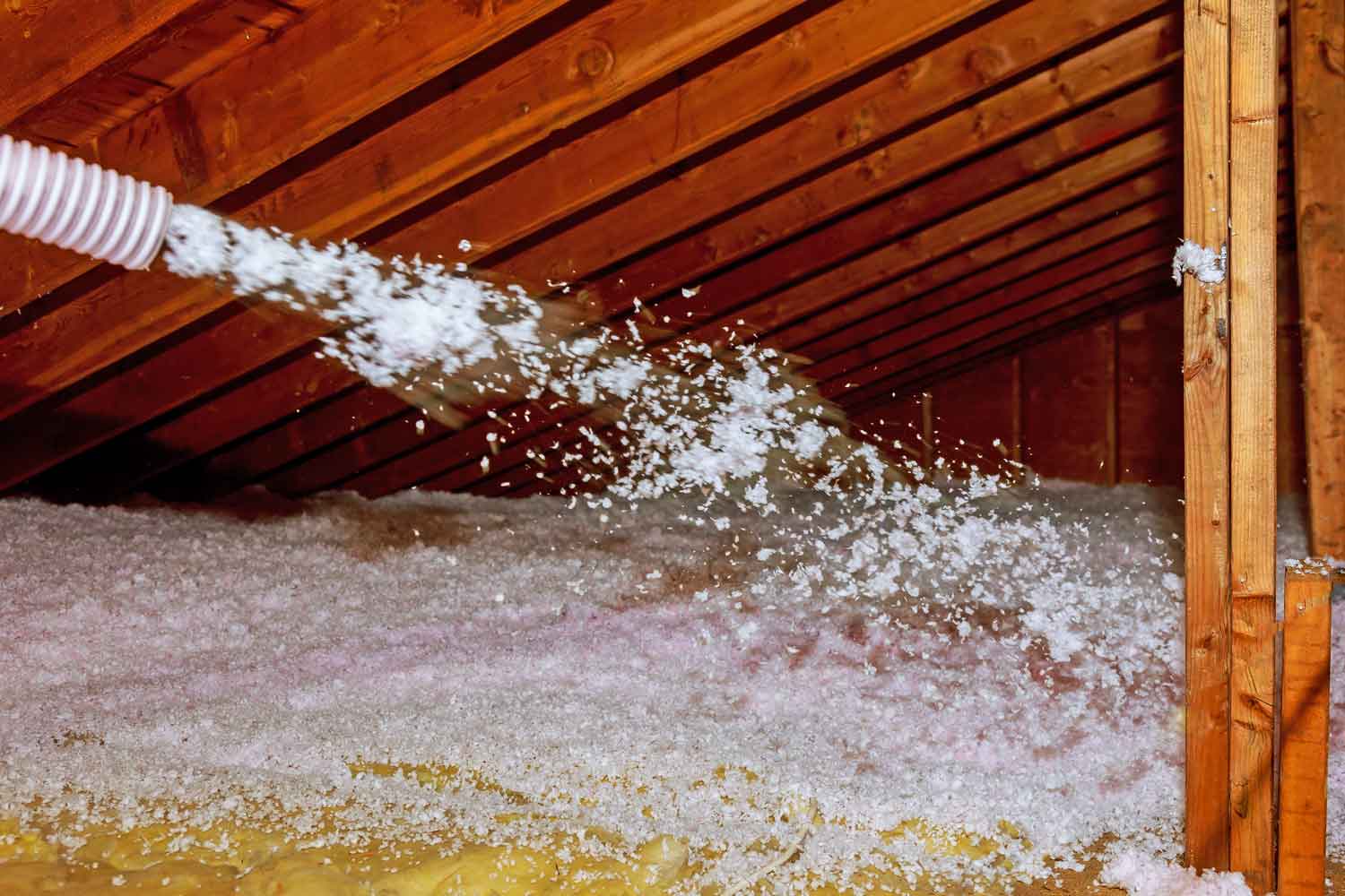 A hose sprays loose-fill insulation between wooden beams in an attic, creating a thick layer of white material on the floor for added thermal protection.