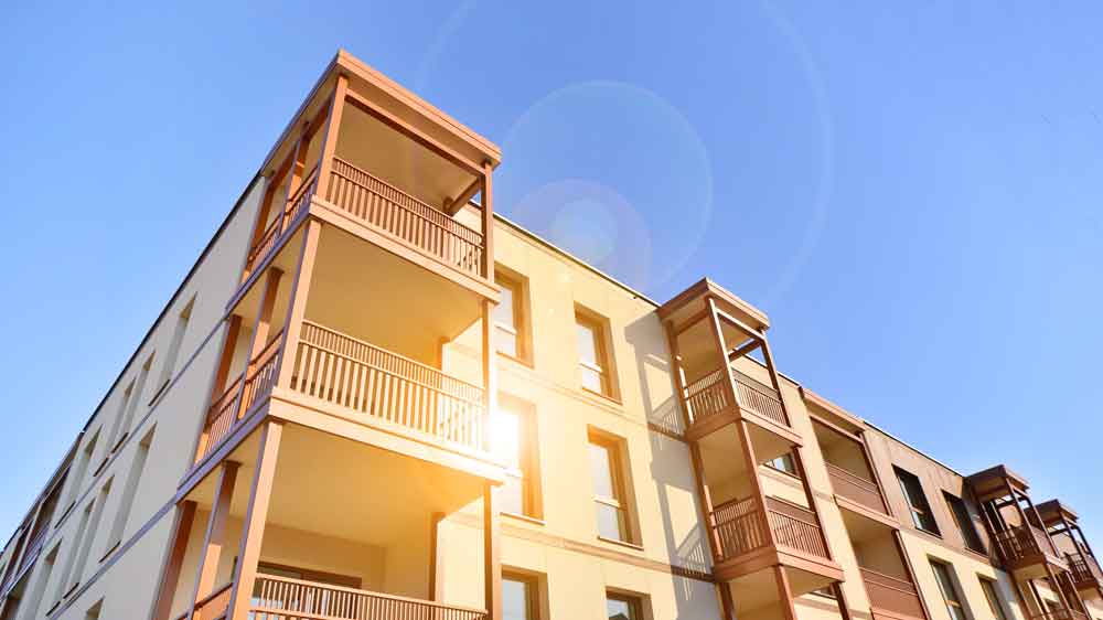 A modern apartment building with multiple balconies and a sun flare against a clear blue sky.