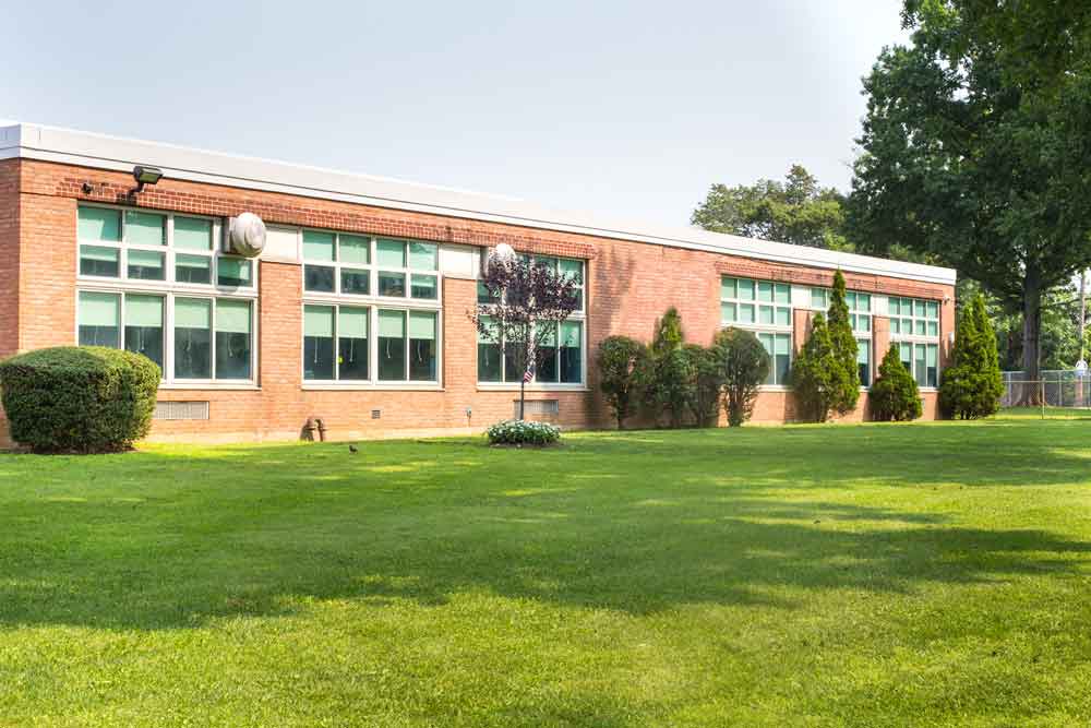 A single-story brick school building with large windows, surrounded by neatly trimmed bushes and trees, stands next to a wide, green grassy lawn on a sunny day.