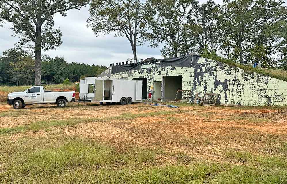 A white truck and a trailer are parked beside a large, weathered concrete bunker with chipped paint, surrounded by grass, dirt, and tall trees under a cloudy sky.