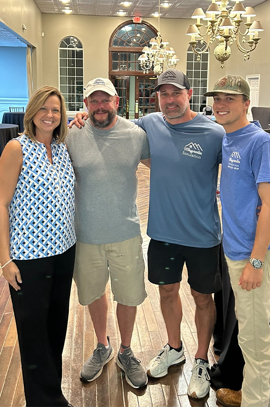 Four people stand together indoors, smiling at the camera. The woman on the left wears a blue patterned sleeveless blouse; the three men next to her wear casual shirts, shorts, and hats in a well-lit room with chandeliers.